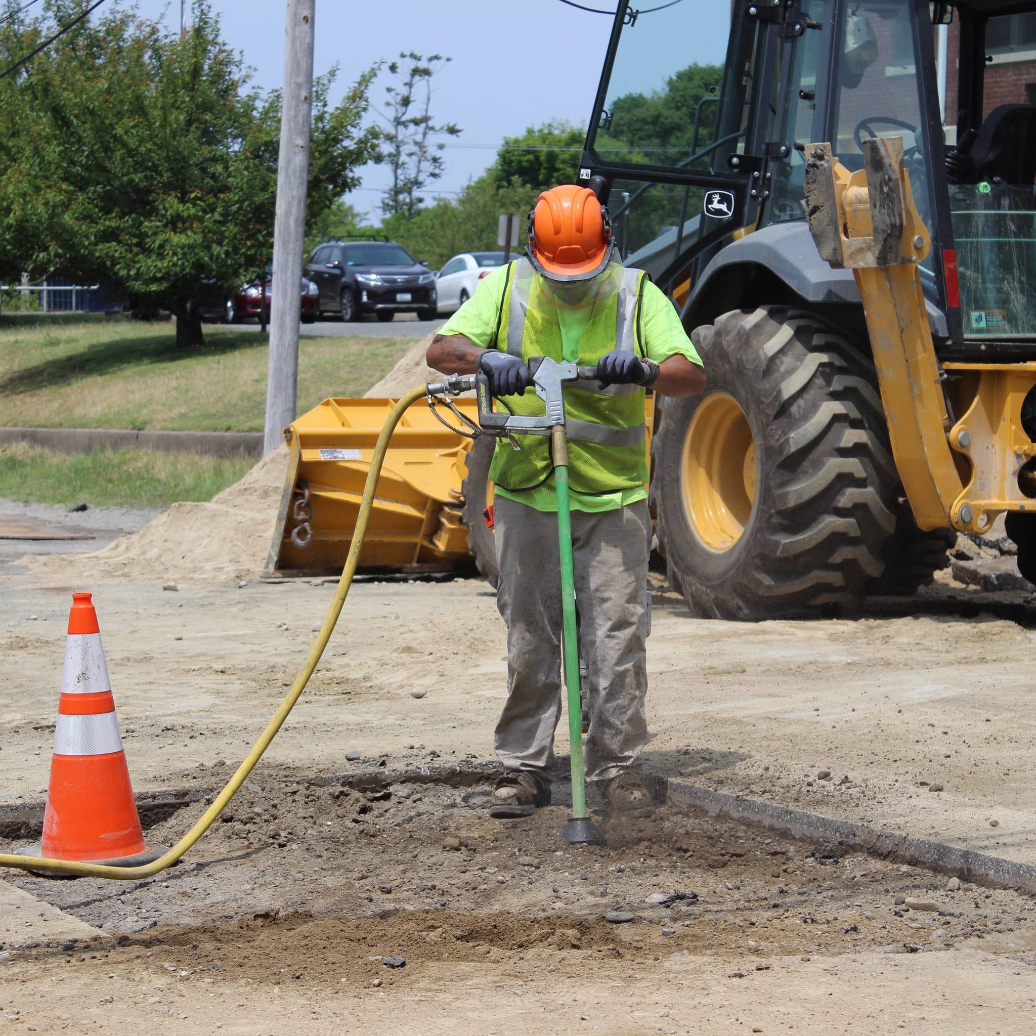 Worker using an AirSpade on a construction site with a vehicle and traffic cone in the background.