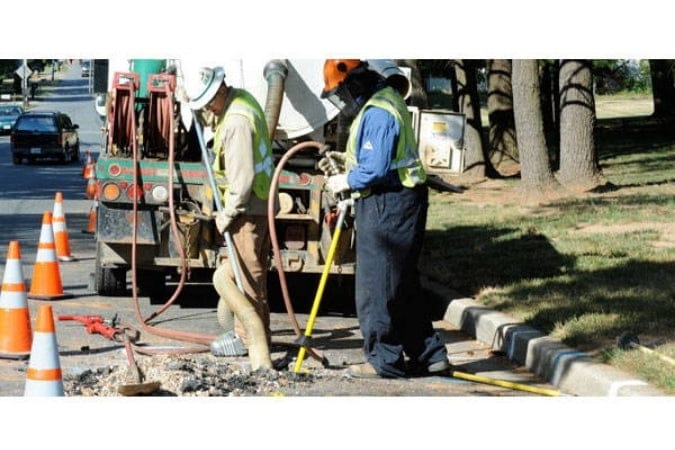 Two workers using an AirSpade on a road with traffic cones and vehicles in the background.