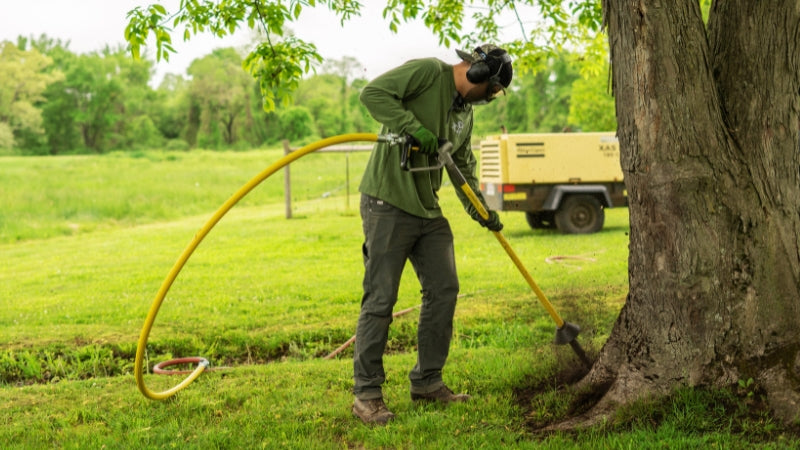 Arborist using an AirSpade near a tree in a grassy area with a truck in the background.