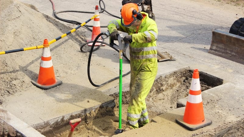 Utility worker in high-visibility clothing using an AirSpade near a hole in the road with traffic cones.